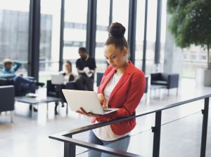 Young afro american woman standing by a railing using laptop with people working in background . Young businesswoman in modern office enviornment.