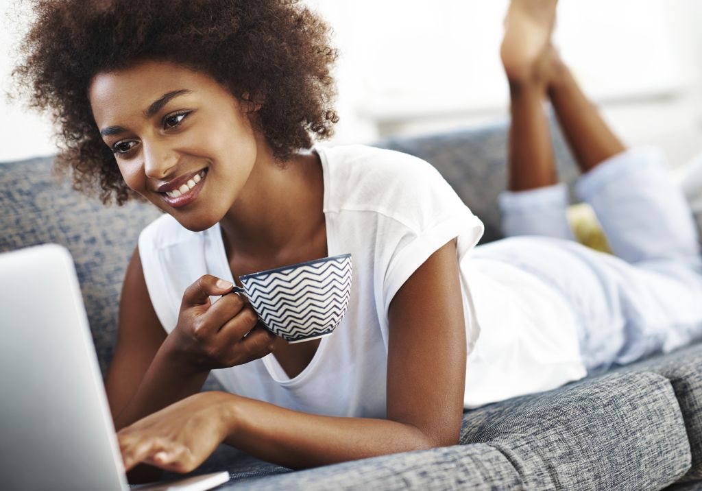 Relaxed ethnic woman lying on her couch with a hot beverage browsing on the internet with her laptop