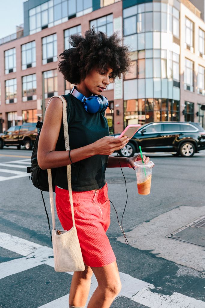 Young woman with headphones and smart phone crossong street in Brooklyn, carying take away drink