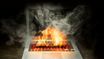 Close up laptop burned with flame and smokes on a wooden desk isolated over black background.