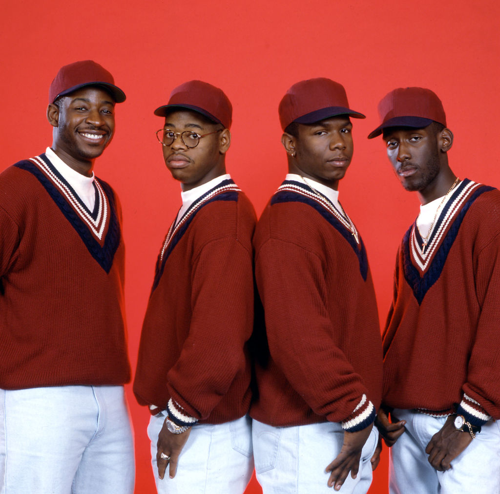Boyz II Men pose against a bright red backdrop in matching red sweaters, white pants, and red baseball caps in a vintage promotional photo from their early career.r