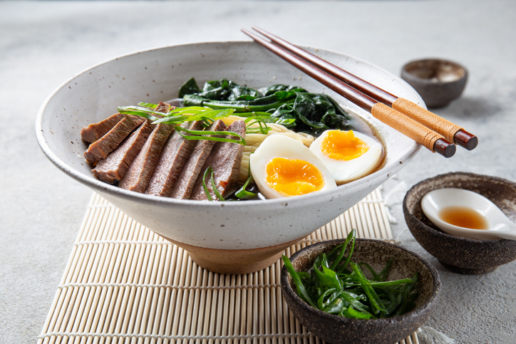 A bowl of ramen with slices of beef, soft-boiled egg halves, and green vegetables. The meal is garnished with green onions