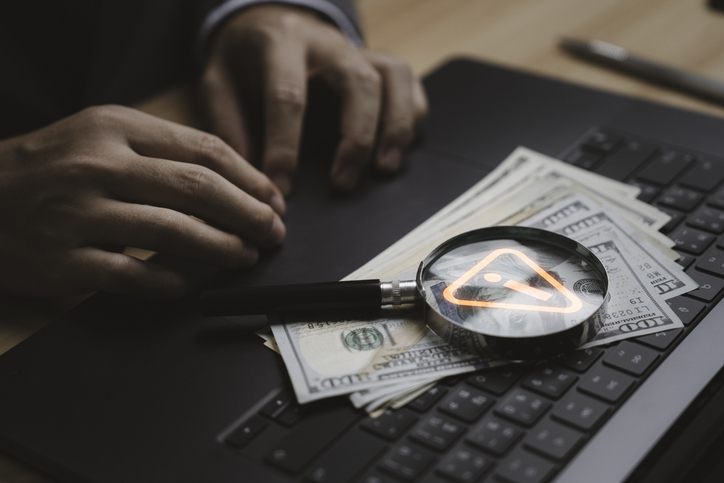 Close up businessman hand working on laptop with dollar bills and caution sign on magnifying glass symbolizing financial scammer money fraud risk online security cybercrime and digital economy safety