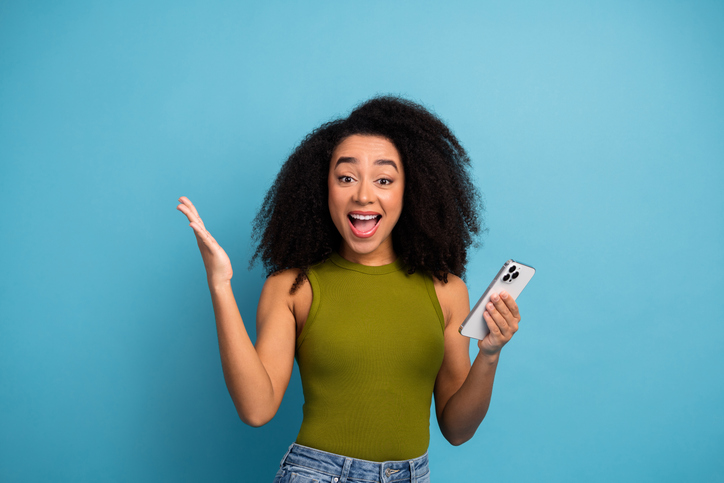 Happy woman with a smartphone excitedly gesturing against a blue background wearing stylish casual fashion attire