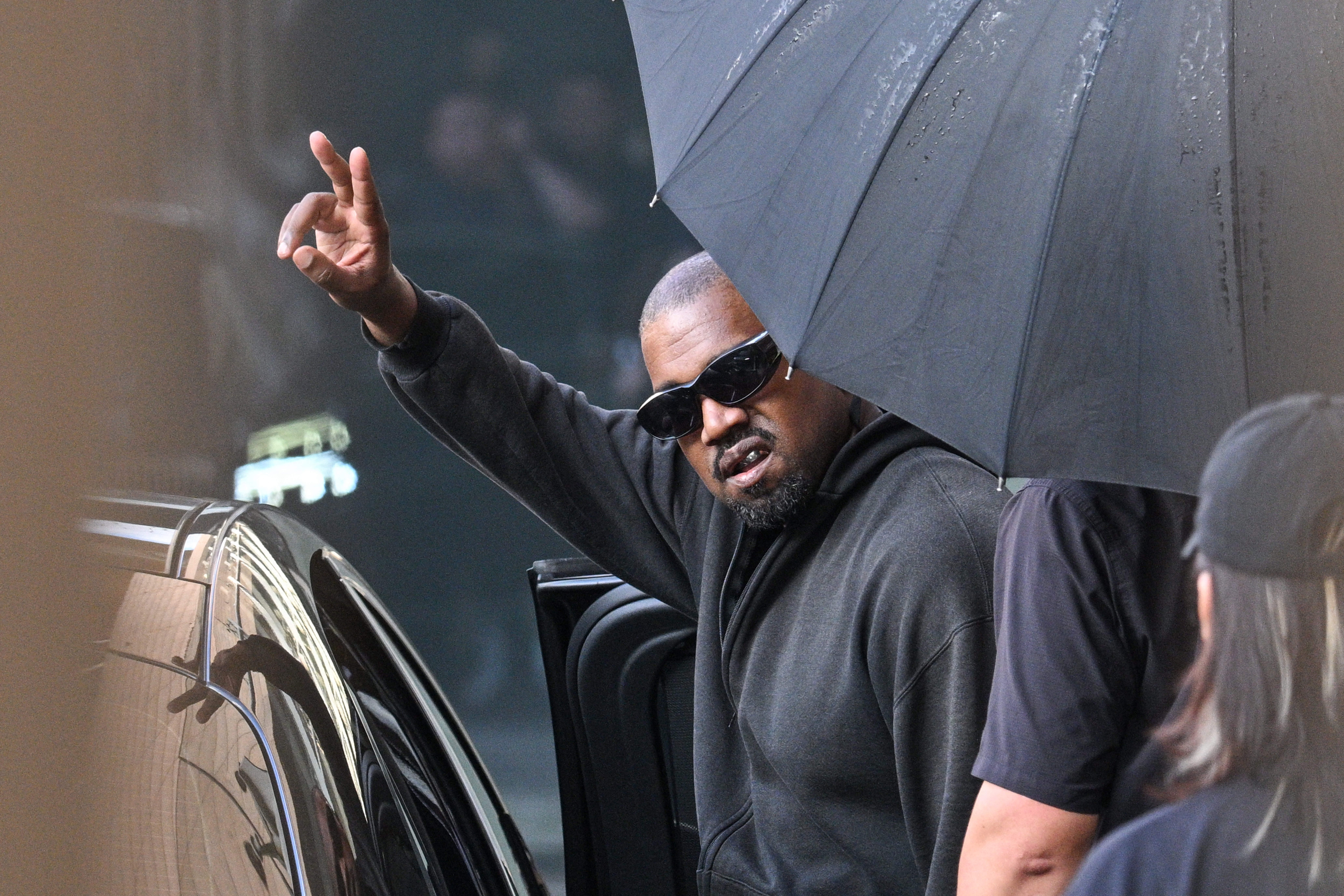 A man wearing sunglasses and a black hoodie gesturing with his hand while holding an umbrella in front of a car.