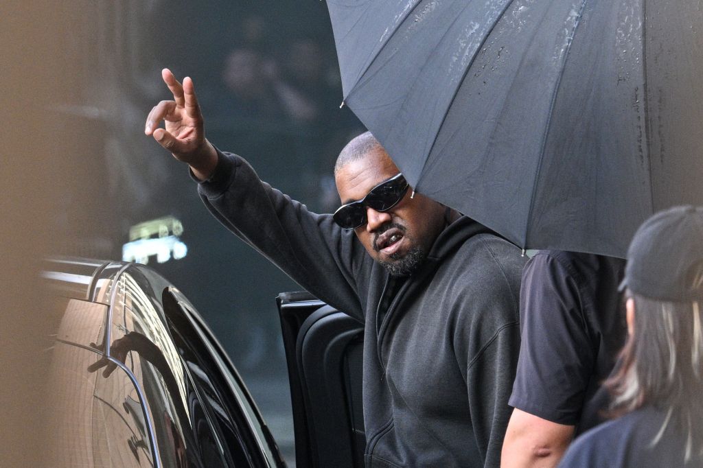A man wearing sunglasses and a black hoodie gesturing with his hand while holding an umbrella in front of a car.