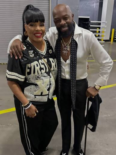 Two people, a woman and a man, standing together and embracing. The woman is wearing a jersey with the text "FIRST 48" and the man is wearing a white shirt and necklace.
