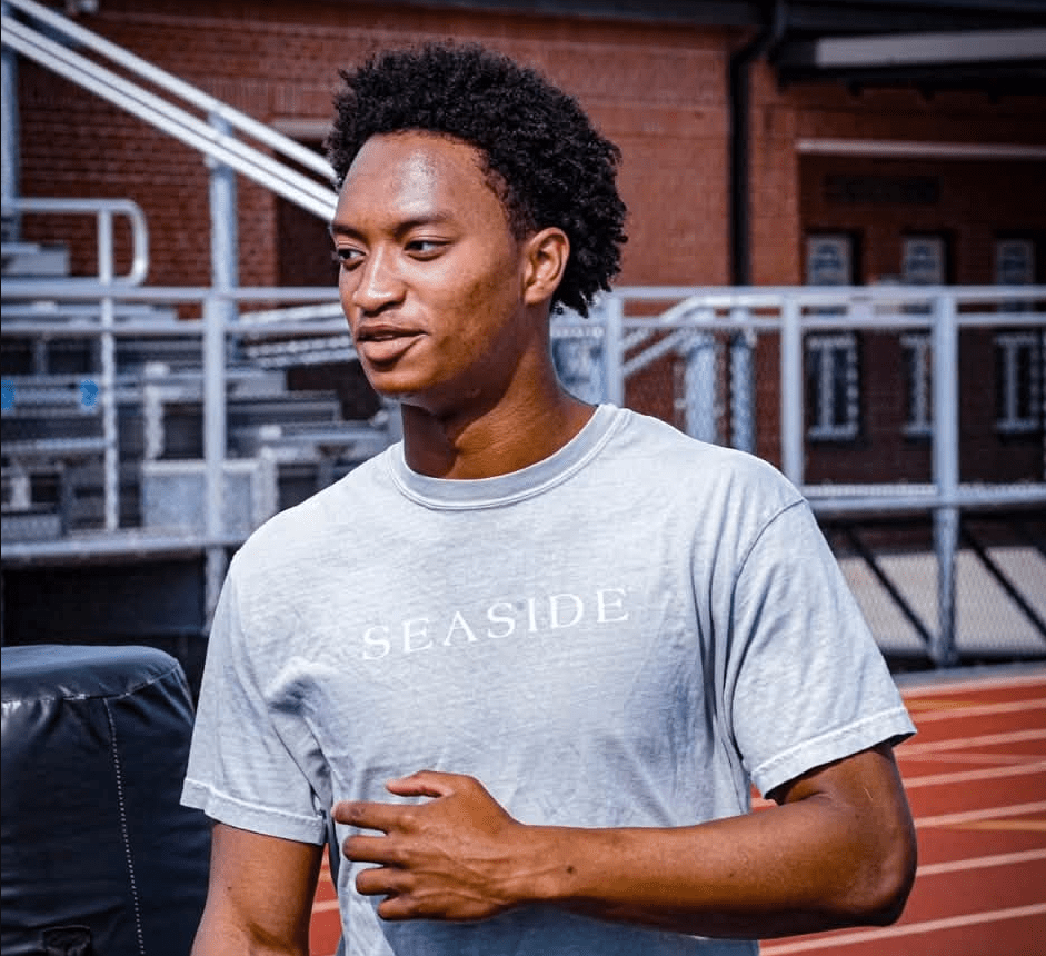 A young Black man wearing a gray "Seaside" t-shirt standing in front of a brick building.
