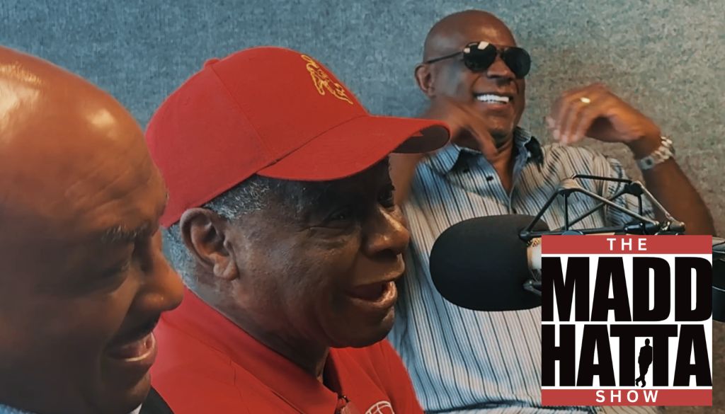 Two men, one wearing a red cap and the other sunglasses, speaking into a microphone on a radio show called "The Madd Hatta Show".