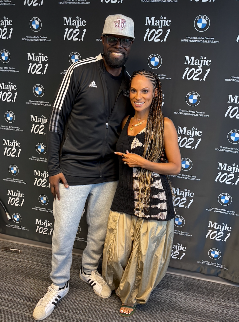 A smiling couple posing in front of a backdrop with "Majic 102.1" branding. The man is wearing a black Adidas tracksuit and a baseball cap, while the woman has long braided hair and is wearing a patterned dress.