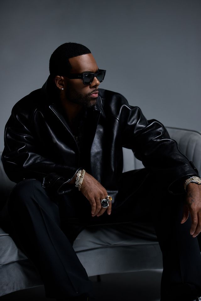 A man wearing a black leather jacket and sunglasses sits on a gray chair, looking pensive.