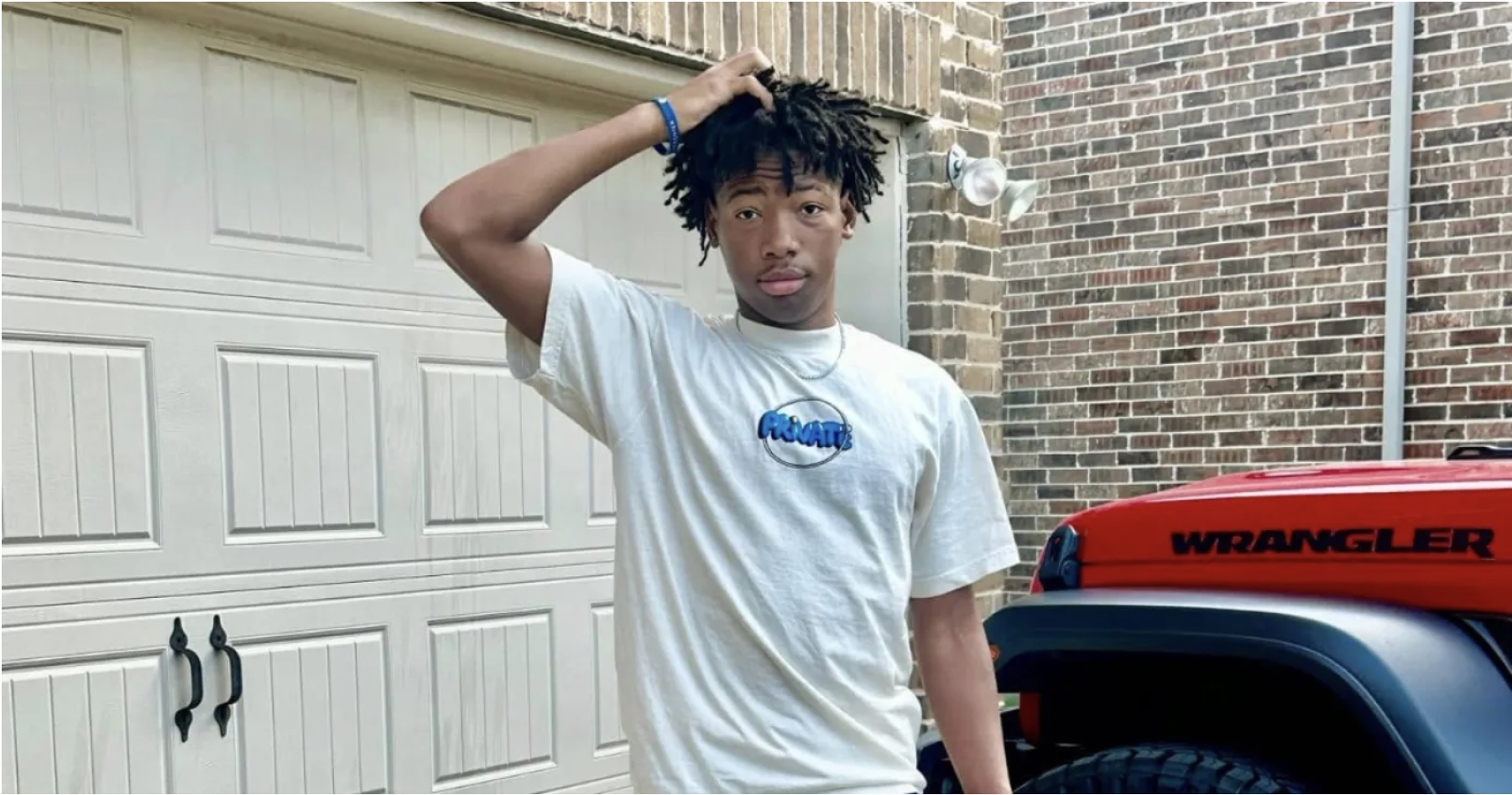 A young man with dreadlocks wearing a white t-shirt with a blue logo, standing in front of a red Jeep Wrangler.