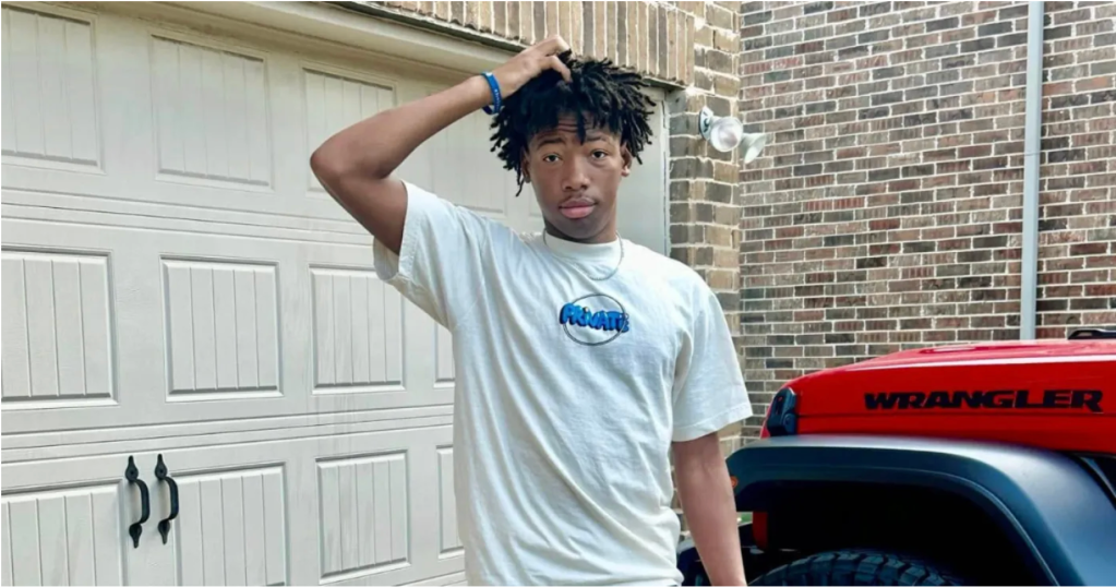 A young man with dreadlocks wearing a white t-shirt with a blue logo, standing in front of a red Jeep Wrangler.