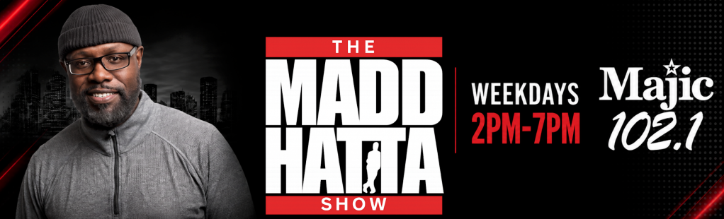 A smiling man with a beard wearing glasses and a gray hooded jacket, standing in front of a city skyline. The text "THE MADD HATTA SHOW" and "WEEKDAYS 2PM-7PM Majic 102.1" is displayed.