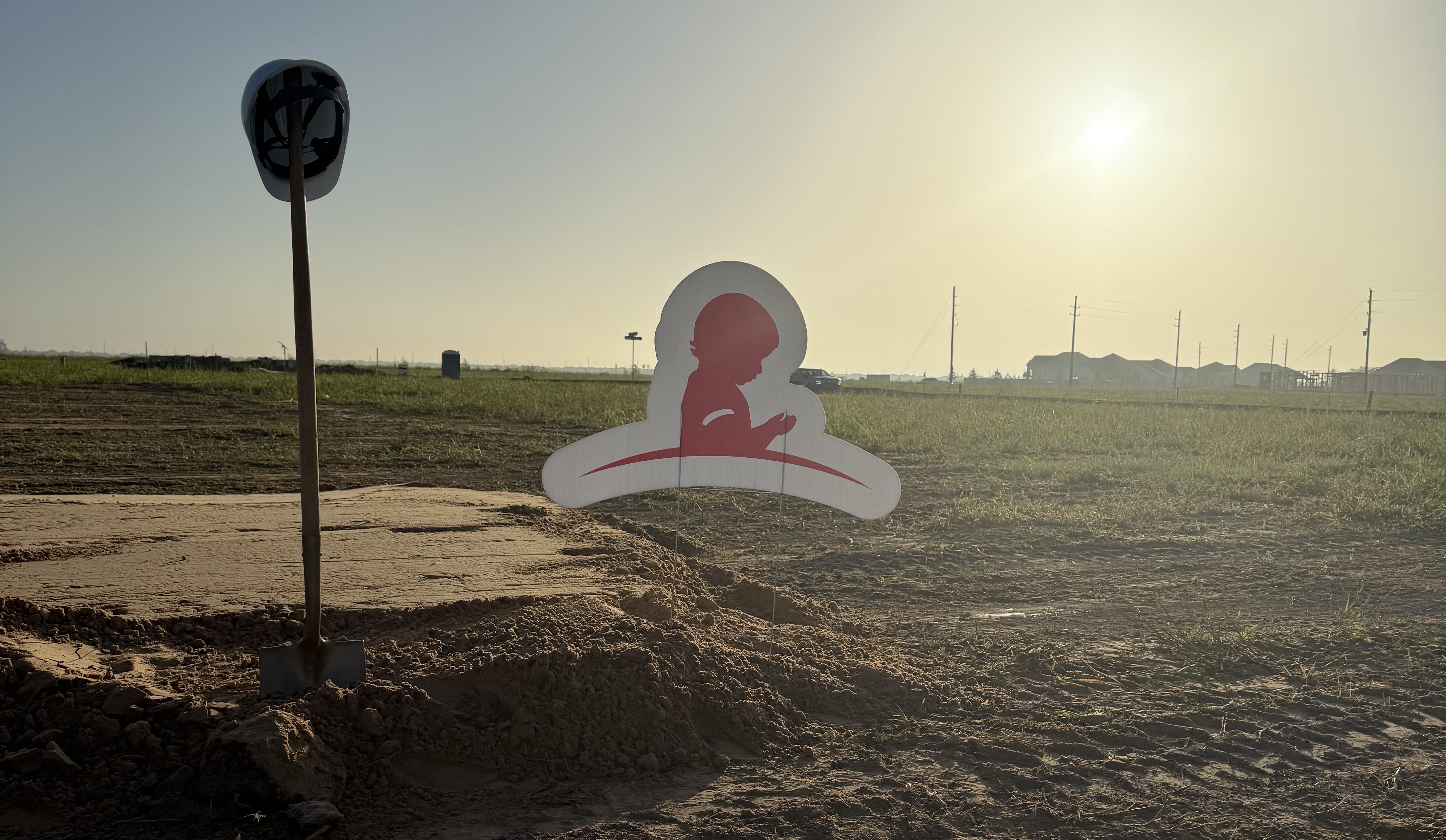 A sunset landscape with a silhouetted figure on a sign in the foreground, against a rural backdrop with industrial structures in the distance.