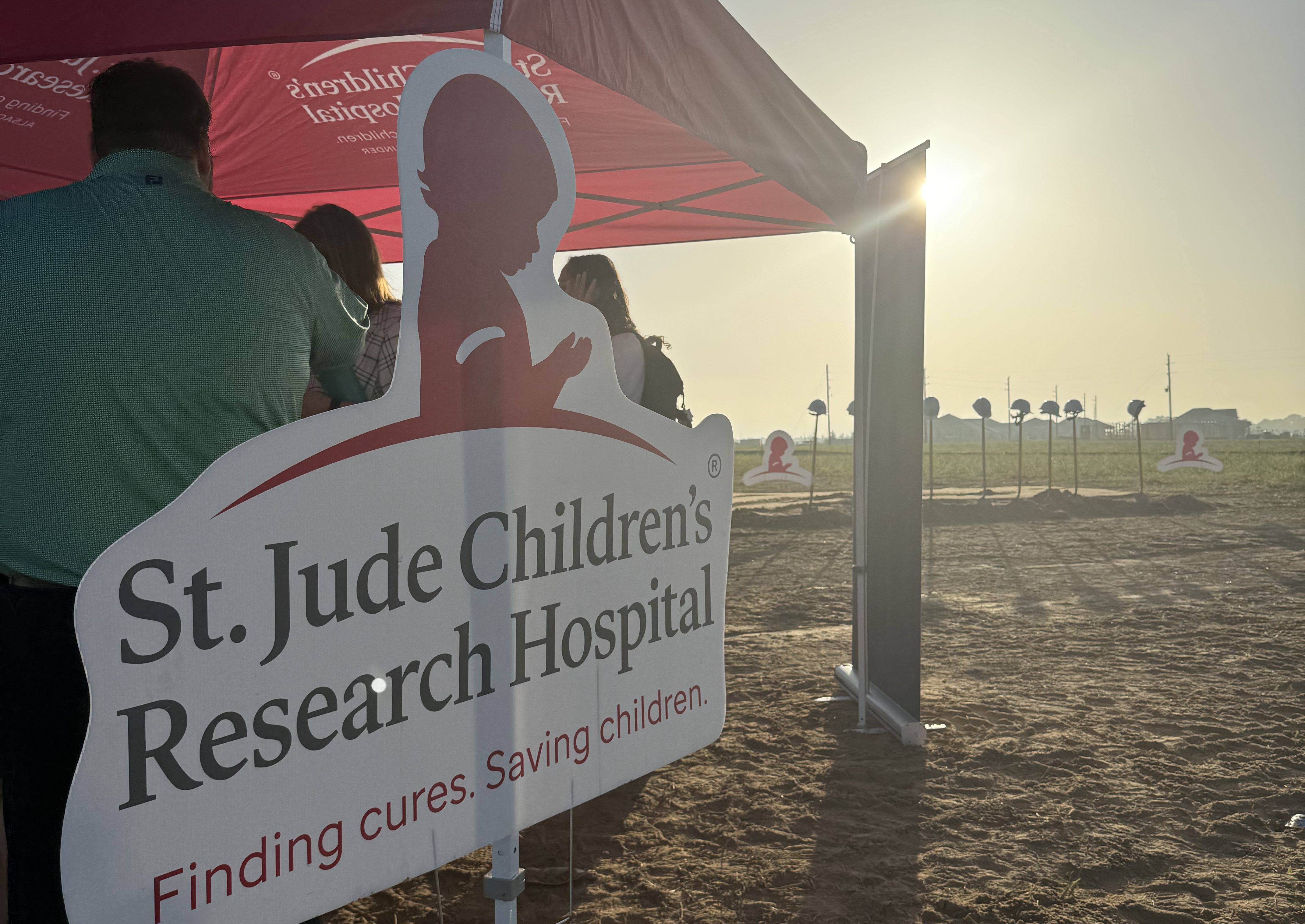 St. Jude Children's Research Hospital sign with silhouette of child against sunset sky, with people in the background.