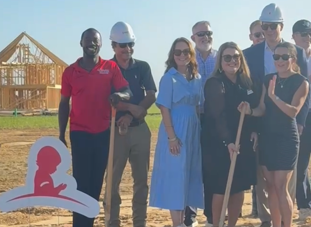 A group of people, some wearing hard hats, standing in front of a partially constructed building. The group includes a mix of men and women.